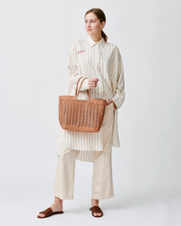 Woman posing in a striped shirt, beige trousers, and brown sandals, holding a Dragon Diffusion hand-woven Window Basket in Natural.