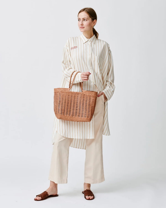Woman posing in a striped shirt, beige trousers, and brown sandals, holding a Dragon Diffusion hand-woven Window Basket in Natural.