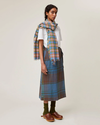 A person wearing a Mois Mont handwoven plaid Scarf No 722 in Nordic Blue, white top, plaid skirt, and brown shoes stands against a neutral background.