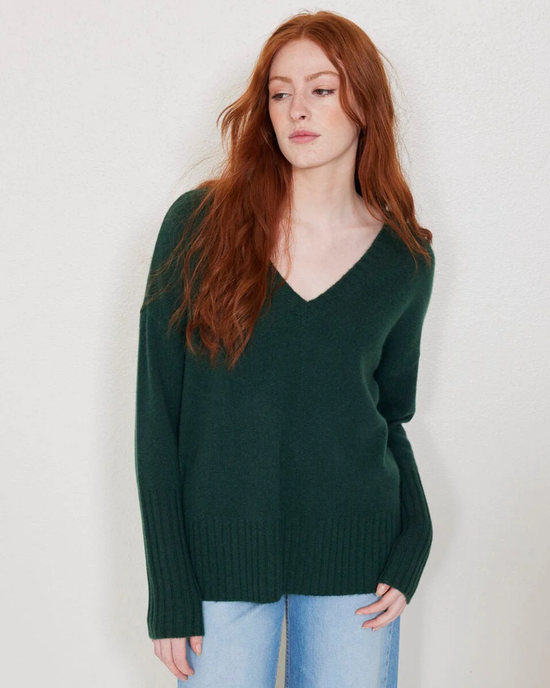 Woman with red hair wearing a green cashmere Ella V Neck in Jade sweater and blue jeans standing against a plain background.