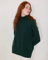 A woman with red hair wearing a green cashmere Elsie Oversize Turtleneck in Jade from Not Monday standing against a white wall.