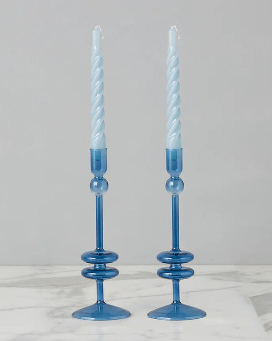 The Sapphire Glass Candlesticks Set of 2 by etúHOME elegantly holds spiral blue taper candles on a sleek marble surface. Crafted from recycled glass, they offer a sustainable touch of beauty to your home decor.