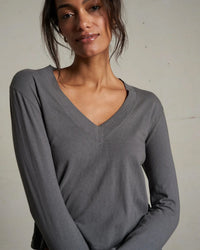 A woman with long brown hair wears the Fiona L/S Recycled V Neck Charcoal top by perfectwhitetee, made from recycled cotton, standing against a plain light background and looking at the camera with a slight smile.