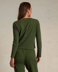 A person with long brown hair stands with their back to the camera, wearing the perfectwhitetee Fiona Long Sleeve Recycled V Neck in Fern Green, paired with matching pants in front of a plain light-colored wall.