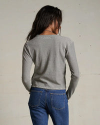 A person with long dark hair, wearing the perfectwhitetee Fiona Stripe Jersey V Neck L/S in Ivory/Black and blue jeans, stands facing away from the camera against a plain, light-colored background.
