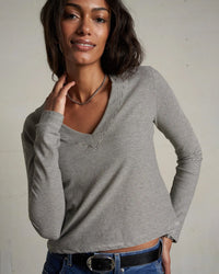 A woman with wavy dark hair wears the perfectwhitetee Fiona Stripe Jersey V Neck L/S in Ivory/Black, paired with blue jeans and a black belt. She smiles slightly, touching her neck while standing against a plain background.