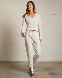 A woman stands indoors wearing the perfectwhitetee Fiona Long Sleeve Recycled V Neck in Silver with matching jogger pants and sneakers, set against an off-white textured wall and concrete floor.