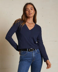 A woman with long brown hair wears the perfectwhitetee Fiona Long Sleeve Recycled V Neck in Navy, paired with blue jeans, a black belt, and a necklace, standing against a plain light-colored wall.