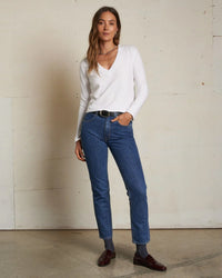 A woman stands indoors by a plain wall, wearing the perfectwhitetee Fiona Long Sleeve Recycled V Neck in White, blue high-waisted jeans with a black belt, maroon loafers, and gray socks, one hand in her pocket.