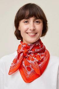 Woman with a short haircut wearing a white top and an orange patterned Mois Mont Poppy Red Bandana No 680 neck scarf, made of 100% Cotton.