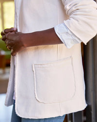 A person wearing the Frank & Eileen Dublin Knit Blazer in Vintage White, featuring a frayed pocket and rolled-up sleeves, stands with hands clasped and turned slightly to the side against a softly blurred background.