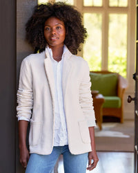 A woman with curly hair stands indoors by a doorway, wearing the Frank & Eileen Dublin Knit Blazer in Vintage White over a white shirt and blue jeans. Sunlight streams in, and a green chair is visible in the background.