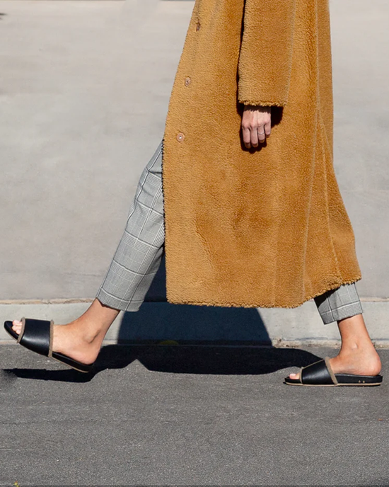 A person walking in plaid pants and a long camel coat paired with the beek. Gallito Shearling in Black/Bronze sandals.