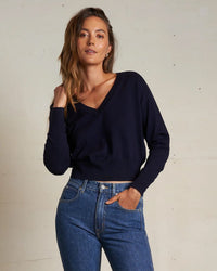 A woman with long brown hair models the Georgina Cashmere V Neck Sweater in Navy by perfectwhitetee and blue jeans, standing with one hand in her pocket and the other touching her hair against a plain light background.