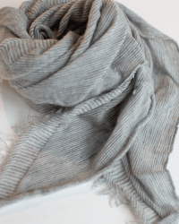 A close-up of Grisal’s Love Mini in Hthr Grey X Milk, a light gray scarf with thin stripes and frayed edges, softly crumpled on a white surface for a gently worn look.