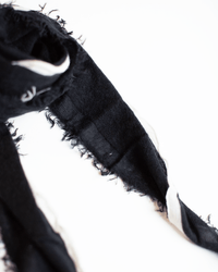 A close-up of the Grisal Rosa Mini in Black X Milk showcases its frayed edges and subtle white detail, laid out on a white surface—an elegant scarf that adds refined style to any outfit.