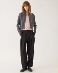A person with wavy hair stands against a plain background, wearing Demylee’s Hallie Cotton Cashmere Cardigan in Dark Heather Grey over a light pink top, loose dark pleated trousers, and black shoes, hands in their pockets.