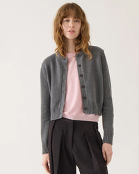 A person with long, wavy light brown hair wears the Demylee Hallie Cotton Cashmere Cardigan in Dark Heather Grey over a pale pink top and black high-waisted trousers, standing against a plain light background.