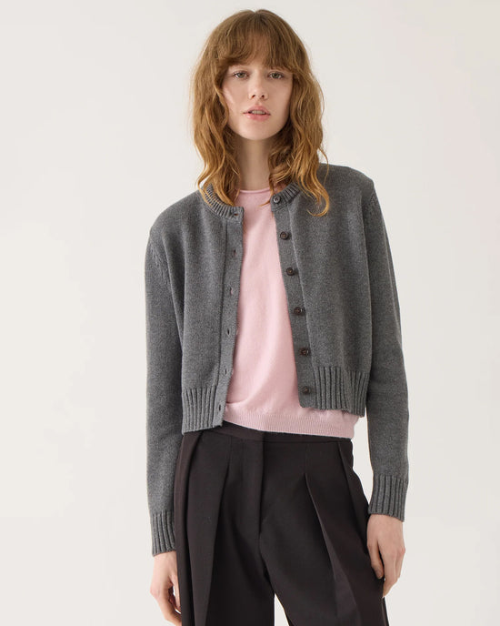 A person with long, wavy light brown hair wears the Demylee Hallie Cotton Cashmere Cardigan in Dark Heather Grey over a pale pink top and black high-waisted trousers, standing against a plain light background.
