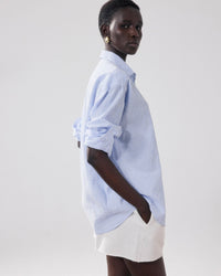 A person wearing the Hartford Charlot Shirt in White & Blue with rolled-up sleeves and white shorts stands sideways, hands in pockets, against a plain light background.