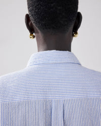 A person with short hair is seen from behind, wearing gold hoop earrings and the Hartford Charlot Shirt in White & Blue.