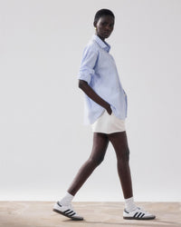 A person wearing a Hartford Charlot Shirt in White & Blue striped cotton seersucker, white shorts, white socks, and white Adidas sneakers with black stripes walks on a light floor against a plain white background.
