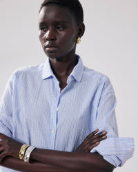 A person with short hair wears Hartford's Charlot Shirt in White & Blue, gold earrings, and silver-gold bangles, standing with arms crossed against a plain light background.