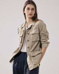 A woman with long brown hair wears Hartford's Vea Jacket in Straw over a white shirt and dark trousers, standing hands-in-pockets against a plain light background. She completes the look with gold hoop earrings.