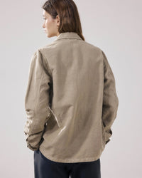 A person with long brown hair stands facing away, wearing the Hartford Vea Jacket in Straw and dark pants, against a plain light background.