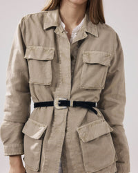 A person wears the Hartford Vea Jacket in Straw—a light beige, military-inspired style with large front pockets and a black waist belt. Made from a soft cotton-linen blend, only the lower half of their face is visible.
