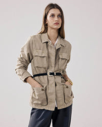 A woman with long brown hair and gold hoop earrings wears the Hartford Vea Jacket in Straw, featuring large front pockets and a black belt at the waist, paired with dark trousers. She stands against a plain background with a neutral expression.