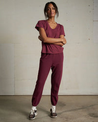 A woman stands against a light wall, wearing the Hendrix Cotton S/S Boxy V Neck Tee in Dark Burgundy by perfectwhitetee with matching joggers, white socks, and brown sneakers. Her arms are crossed as she smiles slightly.