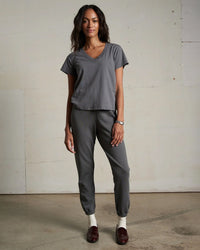 A woman with long wavy hair stands indoors against a plain wall, wearing the perfectwhitetee Hendrix Cotton S/S Boxy V Neck Tee in Charcoal, matching joggers, white socks, and dark brown shoes, with a relaxed expression.