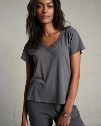 A woman with wavy dark hair models the perfectwhitetee Hendrix Cotton S/S Boxy V Neck Tee in Charcoal, paired with matching gray pants, against a light wall, gazing at the camera with a relaxed expression.