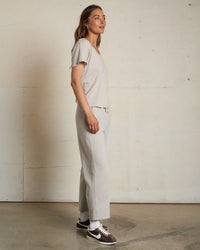 A woman stands sideways against a beige wall, wearing the perfectwhitetee Hendrix Cotton S/S Boxy V Neck Tee in Silver with matching pants, white socks, and black Nike sneakers. She gazes slightly upward with a neutral expression.