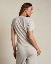 A woman with long brown hair faces away, wearing the perfectwhitetee Hendrix Cotton S/S Boxy V Neck Tee in Silver. She pairs it with matching pants and has a small tattoo on her left arm, standing against a plain light-colored wall.