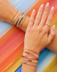 A person's hand adorned with multiple colorful Karen Lazar Design 2MM Sig Bracelets with Pink Sugar Ombre & Yellow Gold bracelets and rings, against a multicolored striped background.