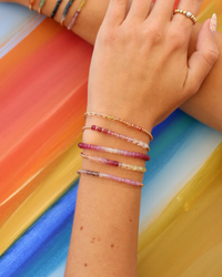 A hand displaying a collection of colorful beaded bracelets, including a 2MM Sig Bracelet with Ruby Ombre & Yellow Gold from Karen Lazar Design, against a rainbow-striped background.