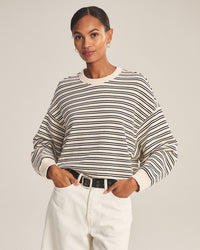 A woman stands against a plain background, wearing the Velvet by Graham & Spencer Isabelle Long Sleeve Crew Neck Top in Natural/Navy, white pants with a black belt, small hoop earrings, and her hair pulled back as she smiles softly.