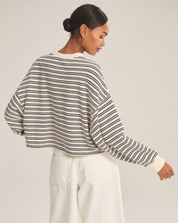 A woman with dark hair in a low bun models the Isabelle Long Sleeve Crew Neck Top in Natural/Navy by Velvet by Graham & Spencer, paired with high-waisted white pants, shown from the back in a neutral studio setting.