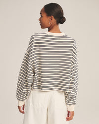 A woman stands facing away, wearing the Velvet by Graham & Spencer Isabelle Long Sleeve Crew Neck Top in Natural/Navy and white high-waisted pants. Her hair is tied in a neat bun against a plain light background—a timeless look.