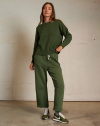 A woman stands indoors wearing the Janet Cotton Sweater in Fern Green by perfectwhitetee, styled with matching wide-leg pants and green Nike sneakers. She has long, straight hair and a relaxed expression, with one hand in her pocket.