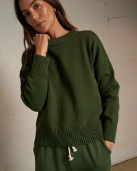 A woman wearing the Janet Cotton Sweater in Fern Green by perfectwhitetee stands indoors, hand on chin and a neutral expression, paired with matching drawstring pants.