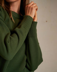 A person wears the perfectwhitetee Janet Cotton Sweater in Fern Green, hands clasped near their face. Their face is partially out of frame, set against a plain, neutral background.