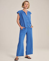 A woman with short blonde hair stands against a plain background, wearing the Janey Ruffle Short Sleeve Button Down in Bluejay by Velvet by Graham & Spencer, paired with matching wide-leg pants and black sandals. Her hands are in her pockets.