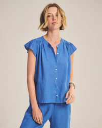 A woman with a short blonde bob wears the Janey Ruffle Short Sleeve Button Down in Bluejay and matching pants from Velvet by Graham & Spencer, accessorized with gold bracelets and rings, standing against a plain light background.