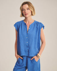 A woman with short blonde hair wears the Janey Ruffle Short Sleeve Button Down in Bluejay by Velvet by Graham & Spencer, paired with matching blue pants. She stands against a plain background, one hand in her pocket and bracelets on her wrist.