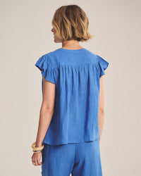 A person with short, light brown hair is seen from the back, wearing Velvet by Graham & Spencer’s Janey Ruffle Short Sleeve Button Down in Bluejay and matching blue pants, accessorized with gold bangles.