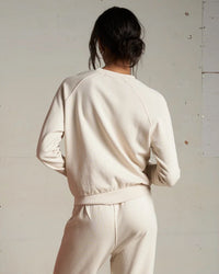 A woman with dark hair in a low bun faces away, wearing the perfectwhitetee Alexi French Terry Sweatshirt in Sugar with matching sweatpants, standing against a neutral, lightly textured wall.