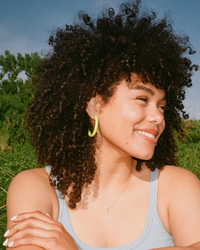 Woman with curly hair smiling in front of a natural backdrop wearing a tank top and B&L sustainably sourced mango wood earrings with 14k Gold Filled Posts.